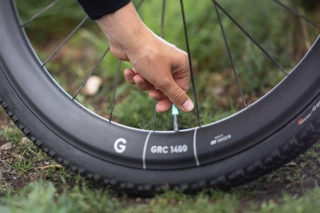 Türkise Ventilkappe eines am Gravelbike-Reifen montierten AERON/TPU Schlauches wird mit der Hand aufgedreht. Im unscharfen Hintergrund befindet sich bemooster Boden.