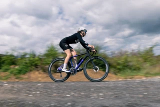 Frau fährt mit ihrem schwarz-lilanen Gravelbike über eine Straße mit bewegungsunscharfem Gestrüpp im Hintergrund.